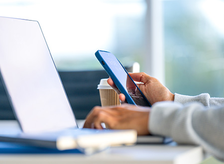 Person holding a phone in one hand and typing on a laptop with the other.