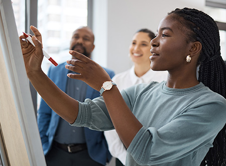 Two business people looking at a white board while one woman writes notes with a marker.