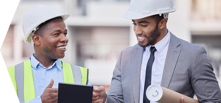 Two men wearing construction hats looking at information on a tablet and holding an architectural drawing.