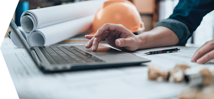 Person leaning on table and typing on a laptop with with architectural plans and hardhat in the background.