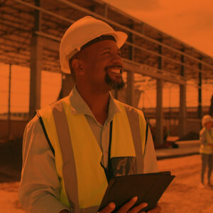 Smiling man in a hardhat and safety vest holding a tablet on a construction site.