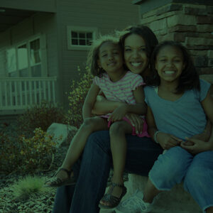 Mother hugging her two young daughters while sitting on the steps in front of their home.