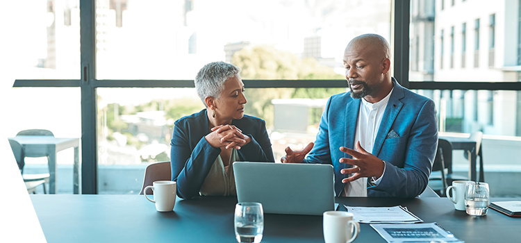 Man and a woman sitting at an office conference table discussing information on a laptop.