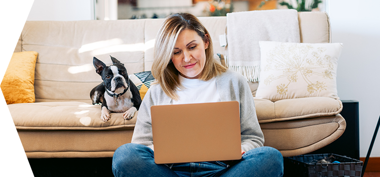 Woman leaning against her couch and typing on a laptop while her dog watches.