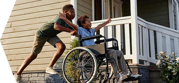 One child pushes another who is in a wheelchair and holds their arms out with joy.